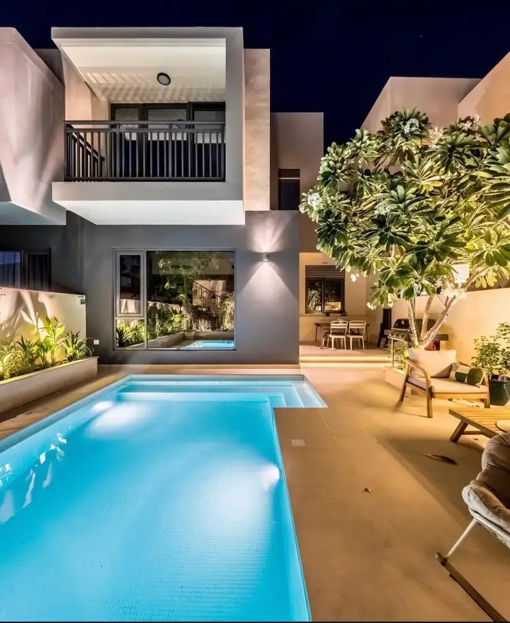Luxury modern pool design at night in Dubai showcasing a glowing turquoise swimming pool framed by light porcelain paving, soft garden lighting, tropical planting, and minimalist white villa architecture — a contemporary outdoor living space crafted by British-owned Reshape Landscaping.