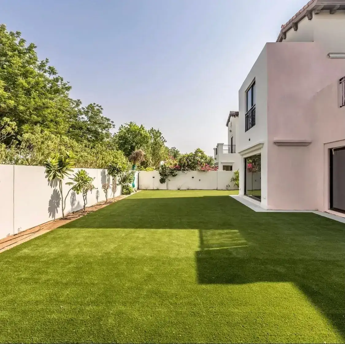 Spacious modern villa garden in Dubai with premium artificial grass, bordered by clean white rendered walls and lush tropical greenery. The design highlights symmetry, open space, and low-maintenance British-style landscaping by Reshape Landscaping Dubai.